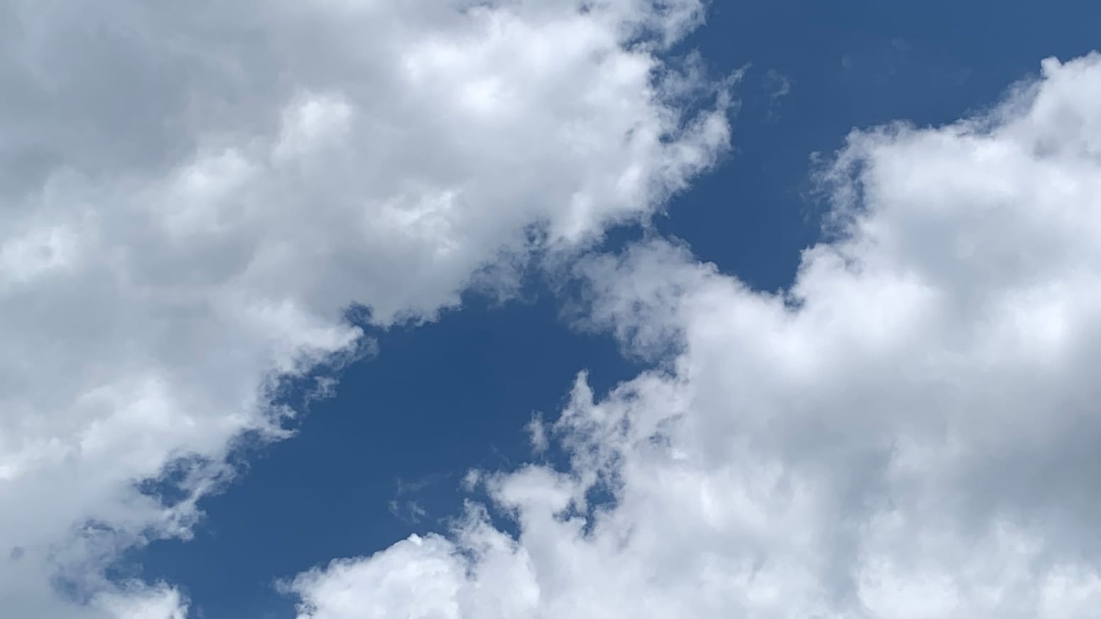 A patch of sky with a sort of gulf or canyon of deep blue running diagonally between dense white clouds, shaded with light gray, which extend in from the top left and bottom right corners of the frame.