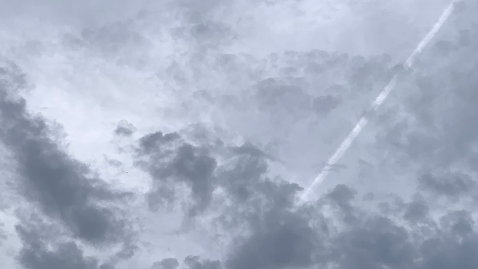 A patch of sky covered in chaotic grays, paler in the background and toward the top but with loose ragged dark gray ones scattered across the bottom of the frame. A straight whitish contrail angles down from near the top right corner, running in front of the lighter gray clouds and disappearing behind the darker ones. 