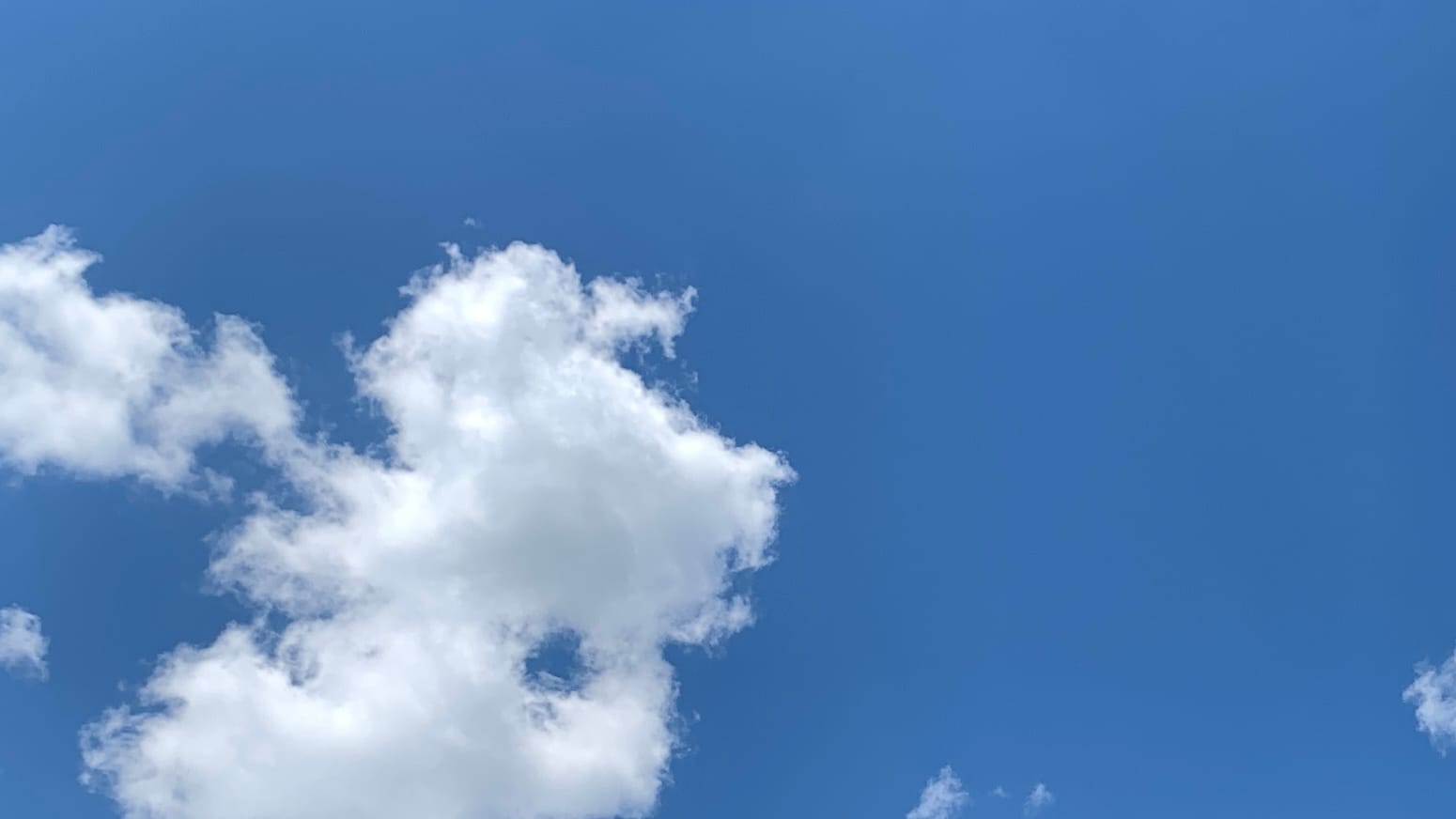 A patch of deep clear blue sky with one cumulus cloud floating it the lower left side of the frame, with a roundish porthole in the cloud showing more blue, a sort of trailing scarf of cloud extending off to the left, and a few small white scraps scattered around the bottom of the photo.. 