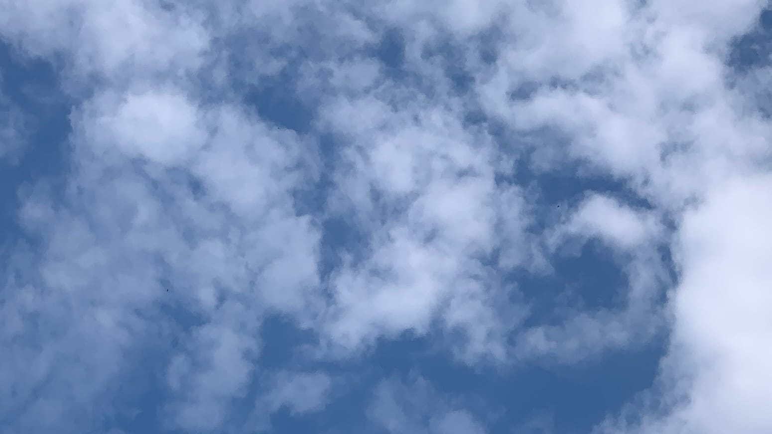 A patch of sky with loose, partly see-through white clouds on it. Toward the lower right part of the frame, where the clouds are a little thicker, a spiral arm of blue reaches in among them, or else a spiral arm of cloud curls around on the blue. . 