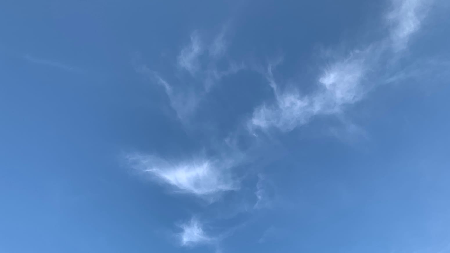 A patch of blue sky with wisps of cloud on it fraying out in multiple directions at once. The wisps are concentrated on a diagonal-ish line running down from the right side of the top of the frame, and vaguely assume the shape of say an iguana windmilling its forelegs. 