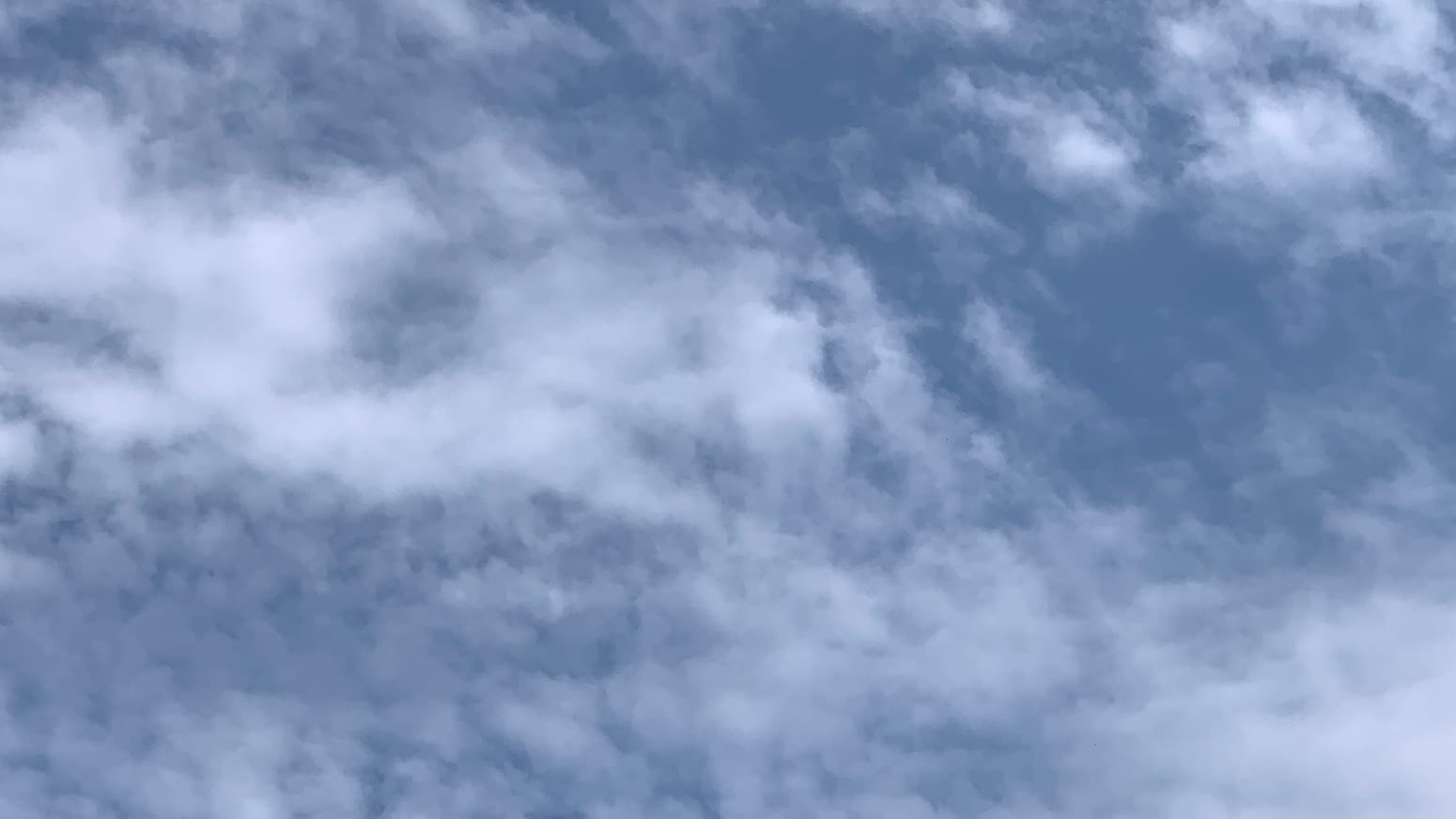 A patch of sky imperfectly covered in thin and loosely attached cloud, thickening to a more solid white in the left middle of the frame and thinning to blue in the right middle. 