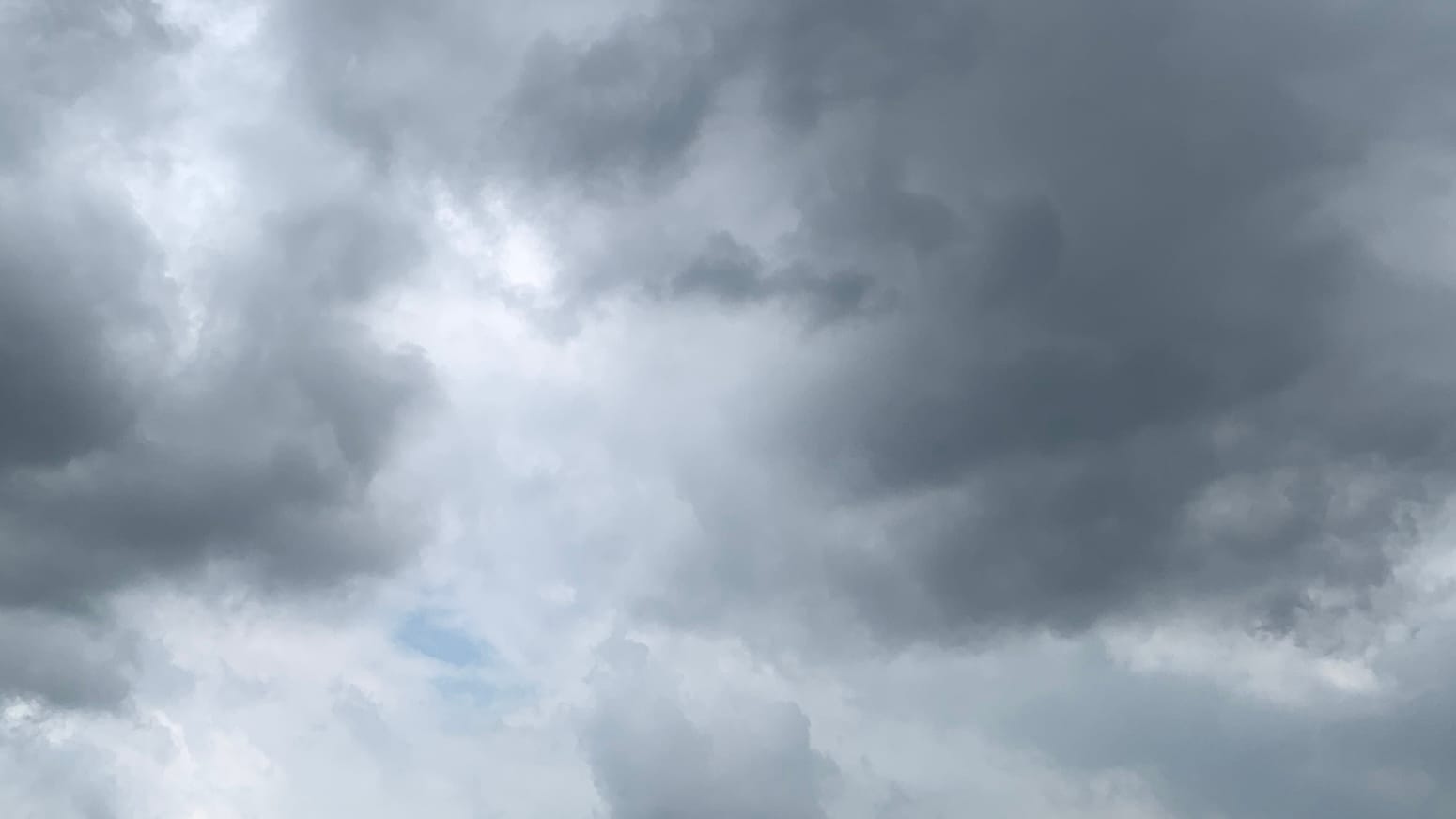A patch of sky with dark gray clouds churning around lighter gray and white clouds. Toward the lower left two little oval holes in the clouds show pale blue through them. 