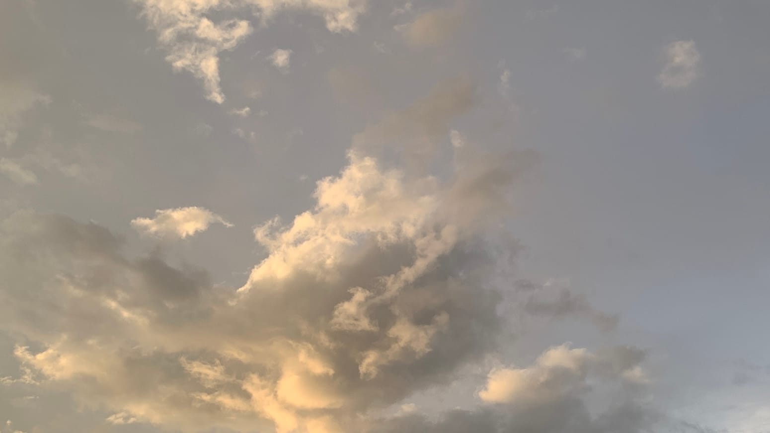 A patch of late-day sky with a jumble of sunlit clouds on it, swollen peach toward the bottom right, whiter toward the center, and overlaid with gray between, with more smoky gray bits floating throughout, all against a background of desaturated blue. 