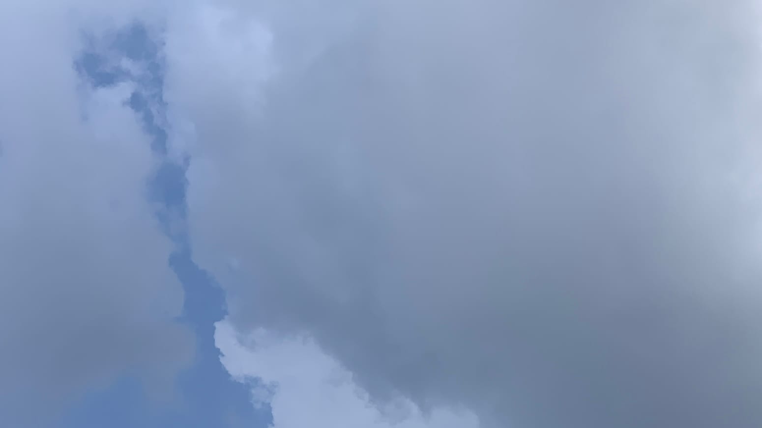 A patch of sky that's mostly gray with a narrow intrusion of blue running near vertically into the frame from the leftward bottom, and with a lighter gray patch to the bottom left side of the blue. The patches of different colors are strangely distinct and flat-looking, like different colors of paint floating in a marbling bath. 