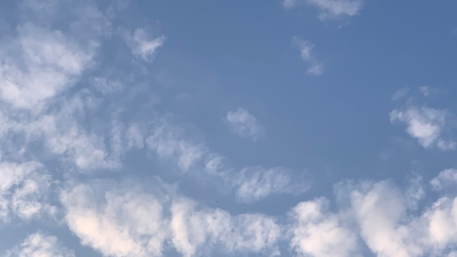 A patch of sky with loose, mostly translucent clouds, faintly tinged with gold, on a blue background. The clouds are curving and are concentrated toward the left and bottom of the frame. 