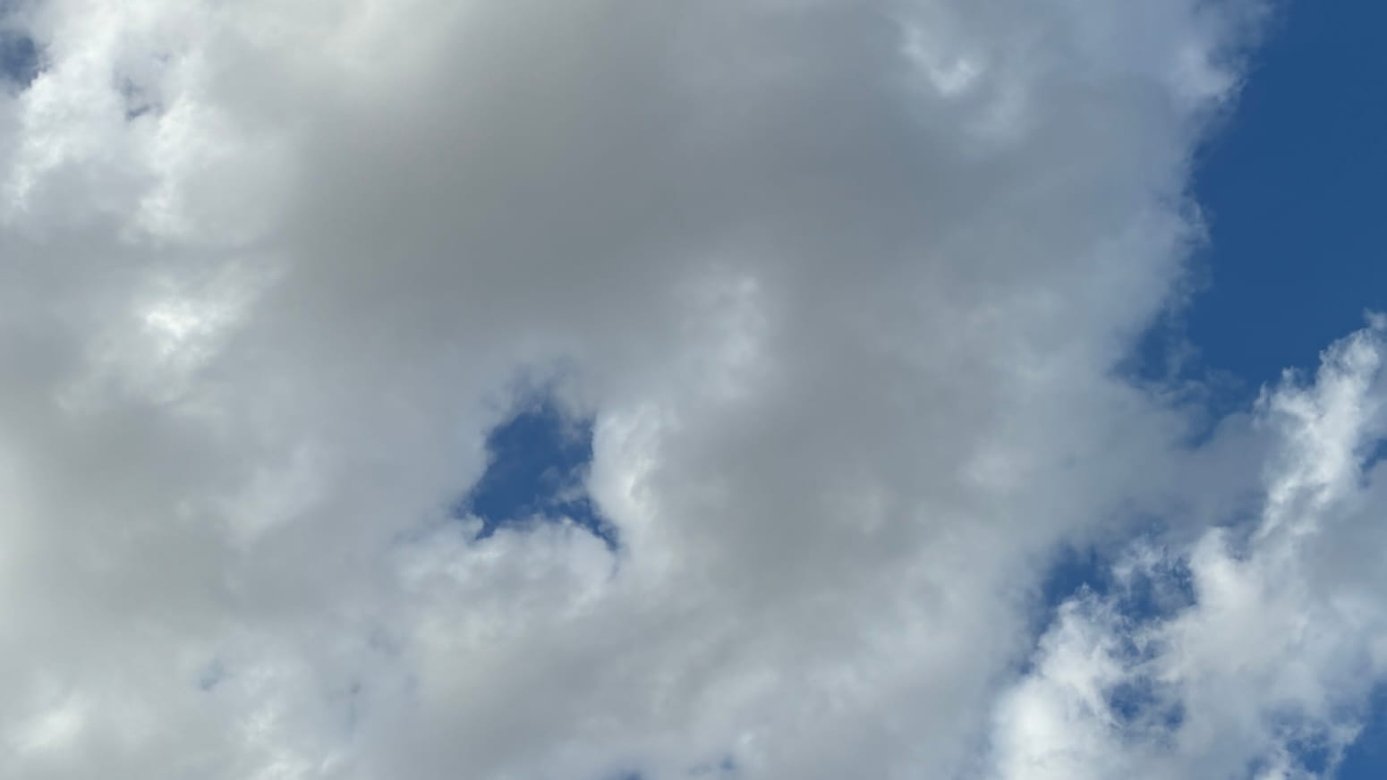 A patch of sky mostly filled with loose-looking white cloud, giving way to deep blue sky around the right edge of the frame. In the left center of the frame is a deep blue hole in the middle of the clouds.