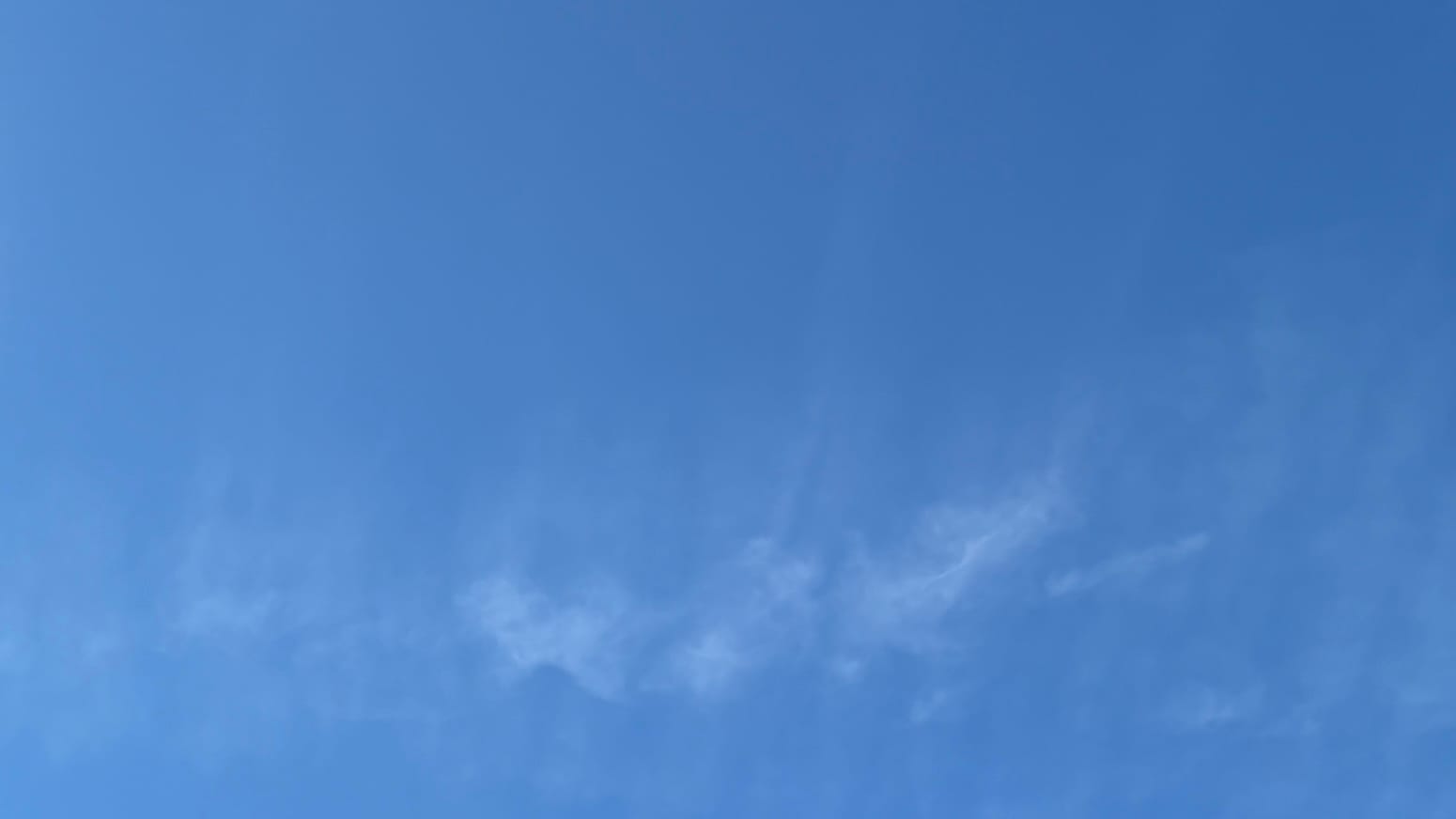 A patch of blue sky with the vanishing, vertically wispy remains of a contrail running across the lower part of the frame. 