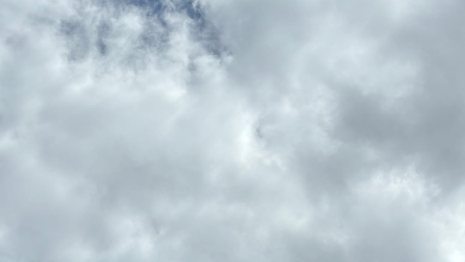 A patch of sky mostly filled with clouds in white shading to gray. A few vertical-ish patches of blue show through a thinning portion of the clouds around the top left. 