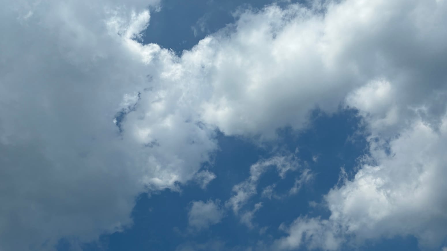 A patch of sky with gray-shaded white clouds on the left and right sides of the frame and a bridge of more cloud connecting those clouds diagonally across a space of blue. 