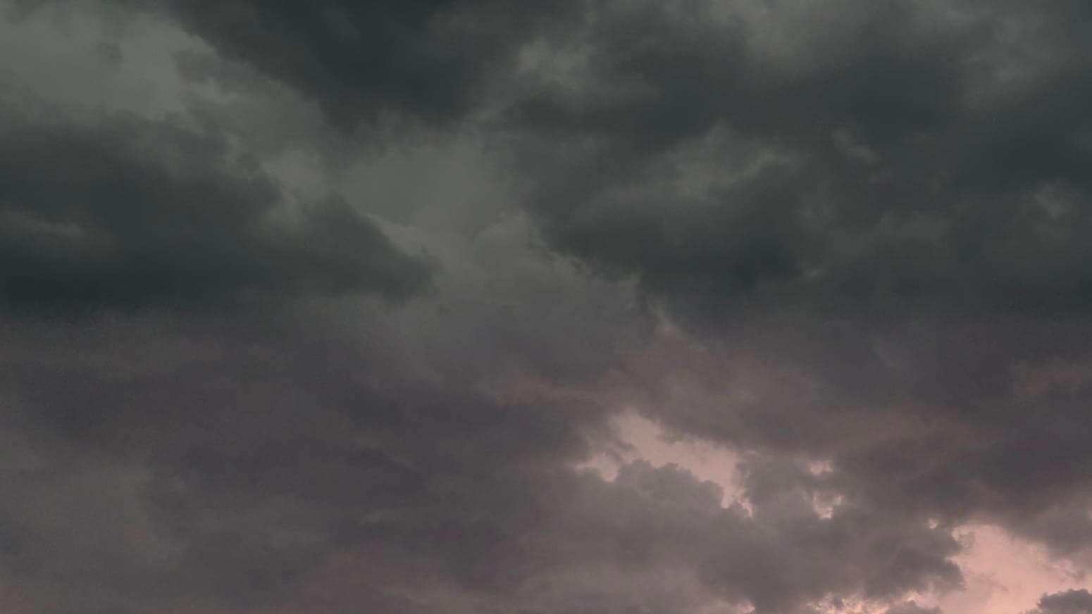 A patch of sky filled with near-black clouds on dark gray clouds. A pink glow from an obstructed lighting bolt rises from the right bottom.