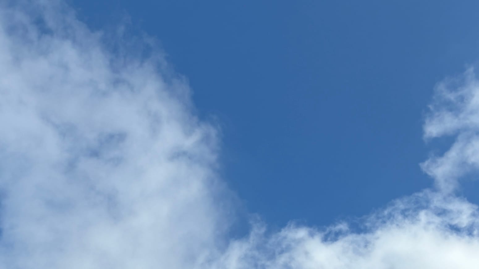 A patch of blue sky framed on the left by a loose, curving white cloud and along the bottom by a denser, straighter white cloud.