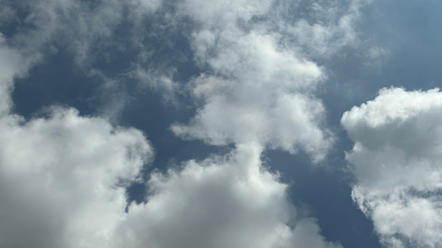 A patch of somewhat faded blue sky with loose, elaborately shaped white clouds overlaid on it. Near the middle of the picture two of the clouds are joined by a short vertical stalk.
