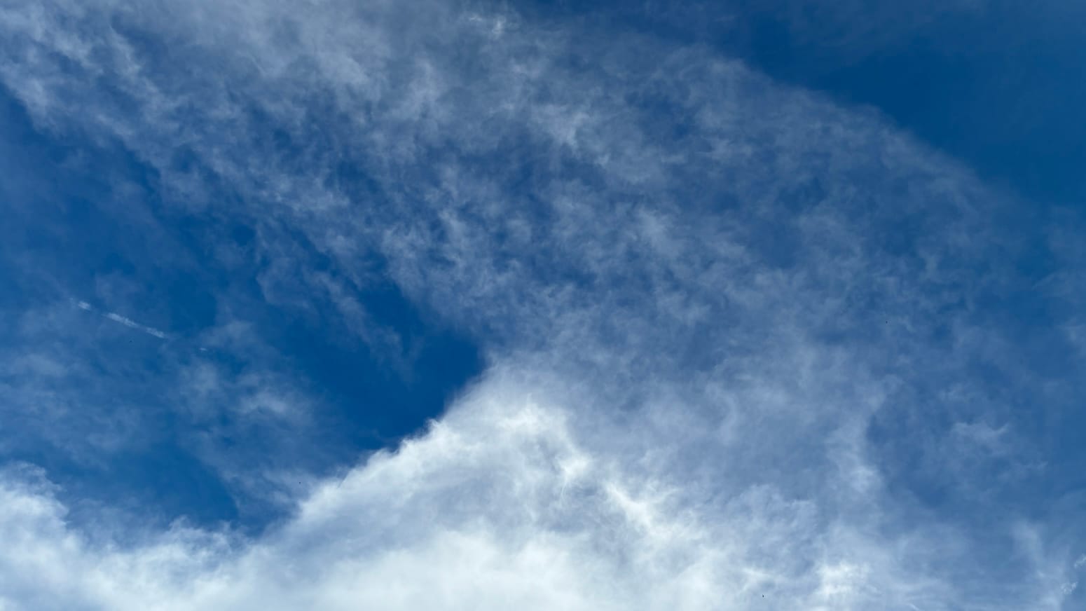 A patch of vivid blue sky with a layer of transparently thin-stretched white clouds overlaid across the upper right half of the frame on a diagonal, which forms one side of a isoceles triangle of denser, luminous white cloud whose base is the bottom of the frame.