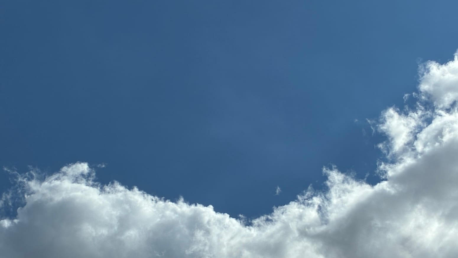 A patch of sky with clear blue above and in the middle and an irregularly zigzagging edge of cloud, with tiny stray flecks of cloud breaking off it, running along the bottom of the frame. 