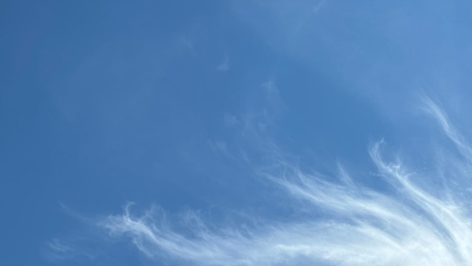 A patch of blue sky, overlaid in places with near-invisible cloud. Wavy tendrils of cloud reach up into the frame from the bottom right corner, as if the rest of a huge cloud-squid were just out of view.  