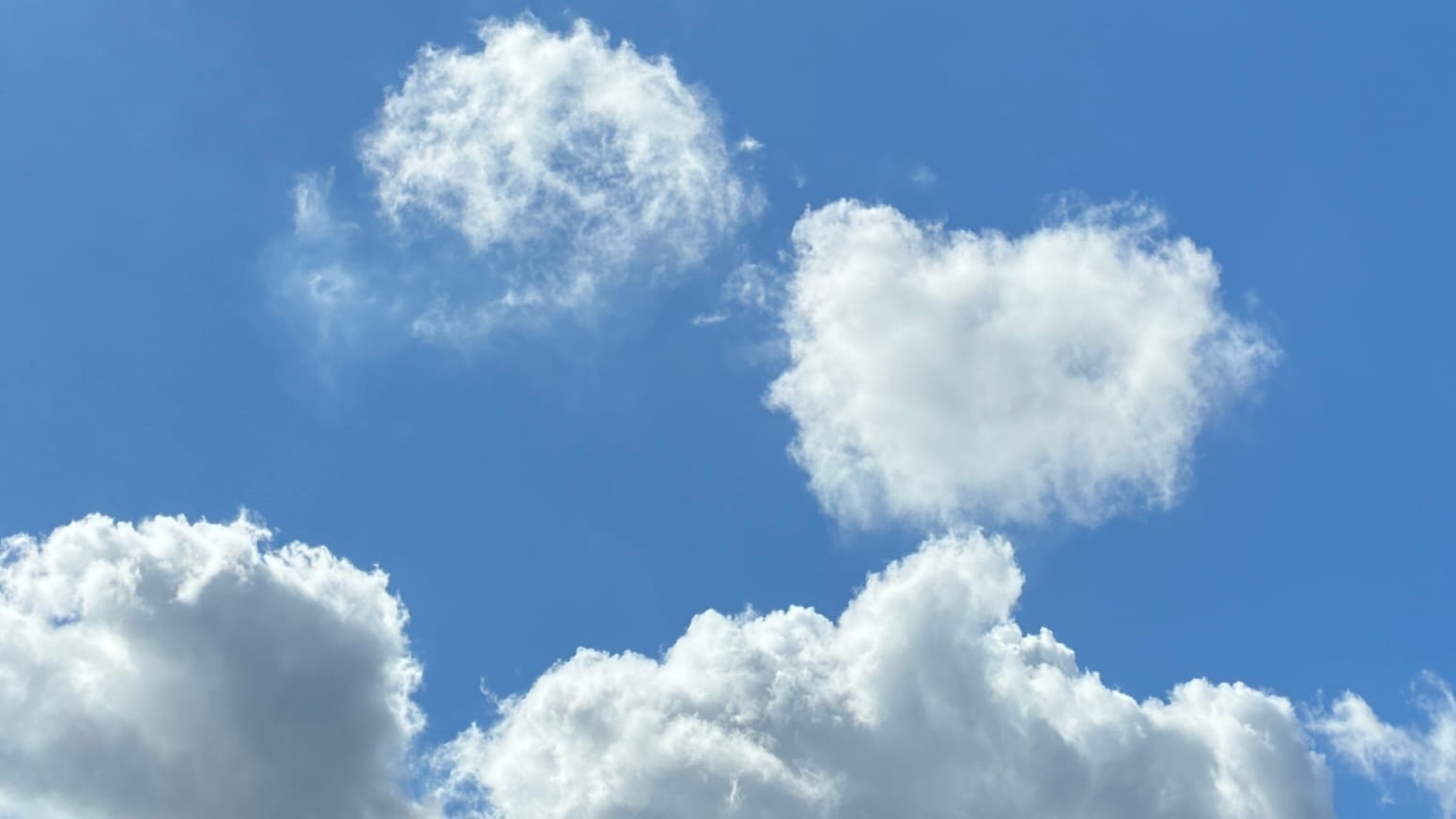 A patch of blue sky with gray-shaded white clouds running along the bottom of the frame and two white clouds floating in the middle, one roughly rectangular and the other a spiral.