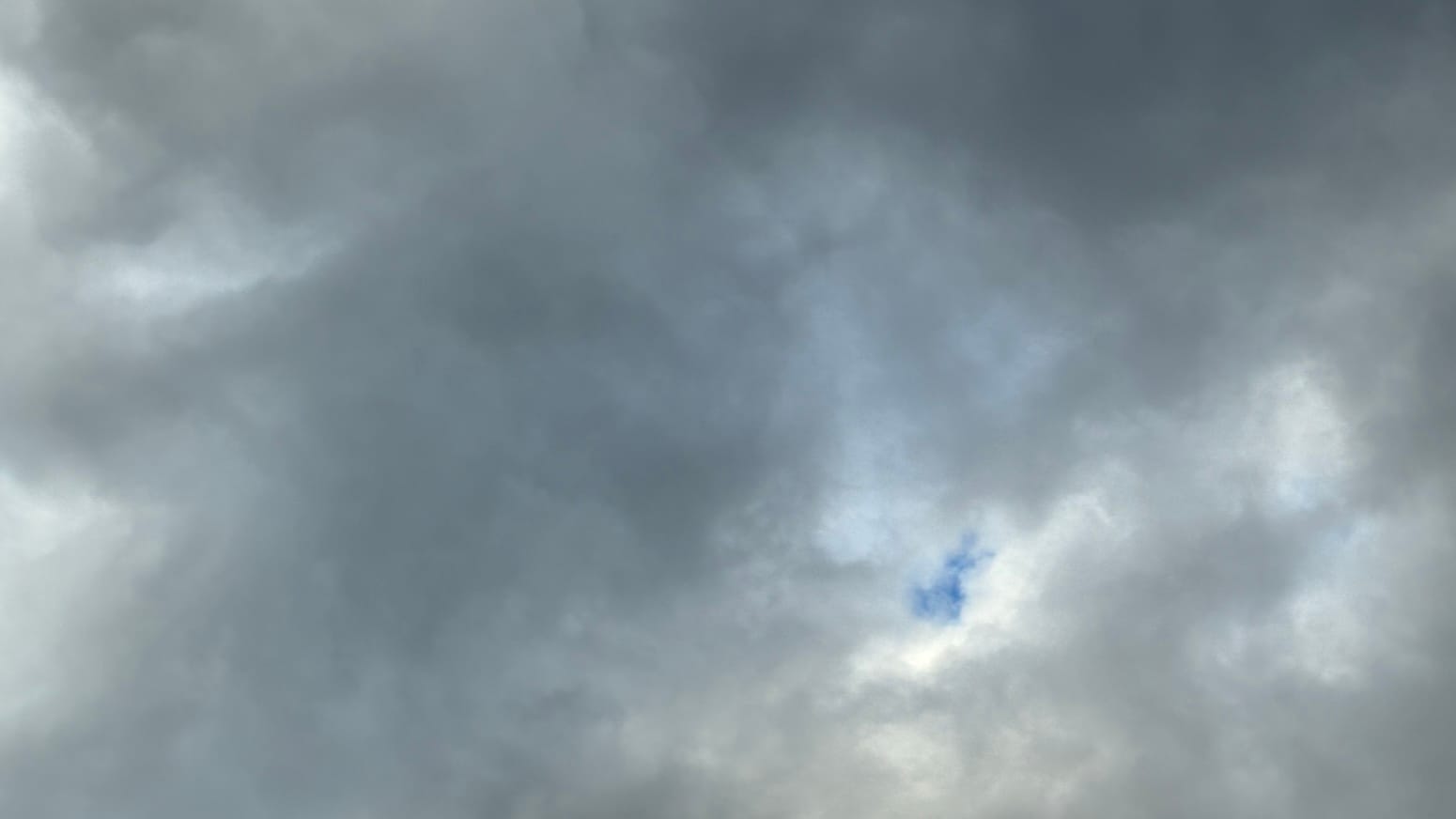 A patch of sky covered in churning clouds in mostly dark and darker gray. In the lower middle right an opening, edged in white, shows a little patch of deep blue sky through it.
