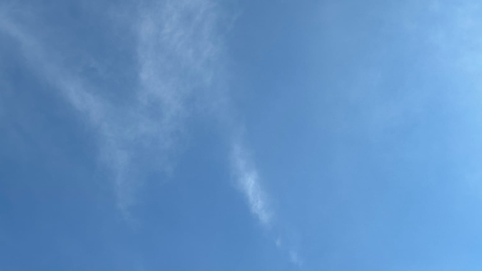 Alt: A patch of somewhat hazy blue sky with a near-transparent cloud resembling a capital letter M in the left side of the frame.