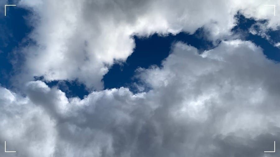 White clouds with grey shadows at top and bottom of frame with a jagged backdrop of deep blue sky