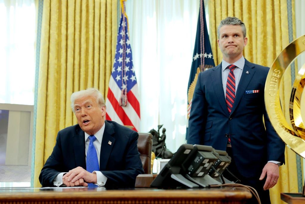 Trump seated semi Oompa Loompa-tinged, happily blathering away as Hegseth stands nearby doing childlike puffy-cheek face