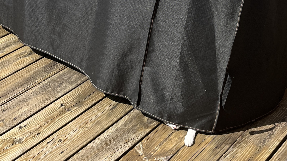 The outdoor deck of my castle where I keep my gigantor smoker/grill with the cover on and there is a paw and the front part of a cat's face sticking out at bottom of the cover.