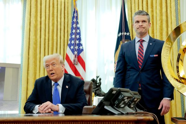 Trump seated semi Oompa Loompa-tinged, happily blathering away as Hegseth stands nearby doing childlike puffy-cheek face