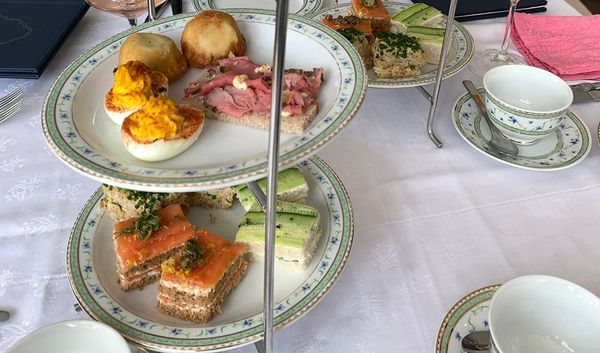 Plates of finger sandwiches and deviled eggs stacked in racks on a table with cups and saucers