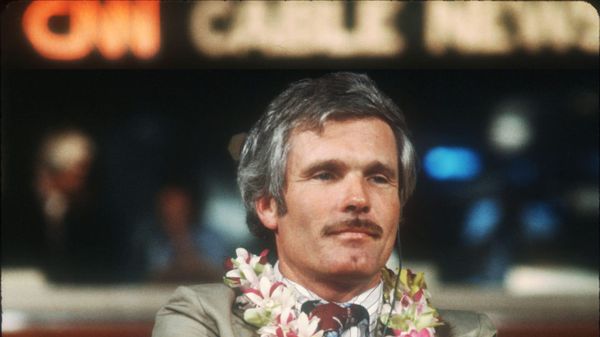 Turner in a tan suit, wearing a lei, sitting in a chair in studio