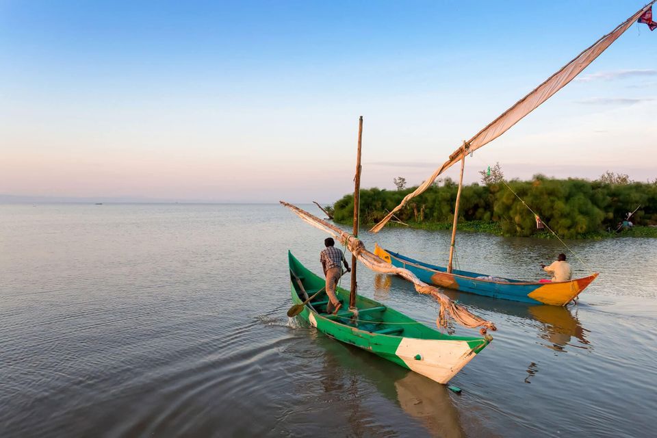 2 small boats with single sails furled each being paddled by a man on calm water