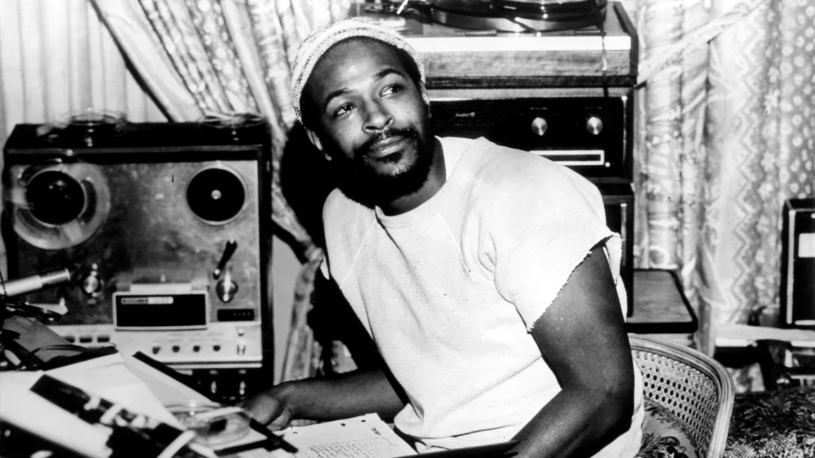 A black and white photo of Marvin Gaye sitting down with a notebook of lyrics in his studio. Photo by Gems/Redferns.