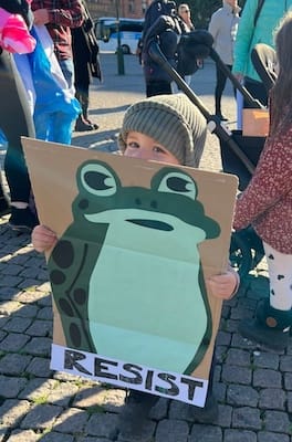 Photo of a child in a green hat peaking over a cardboard sign showing a frog and the word "resist"