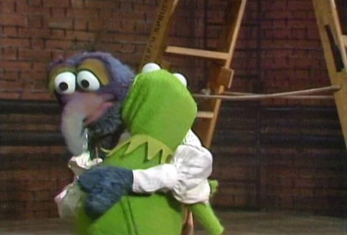 Gonzo hugging Kermit on an empty stage, with a brick wall and a ladder in the background.