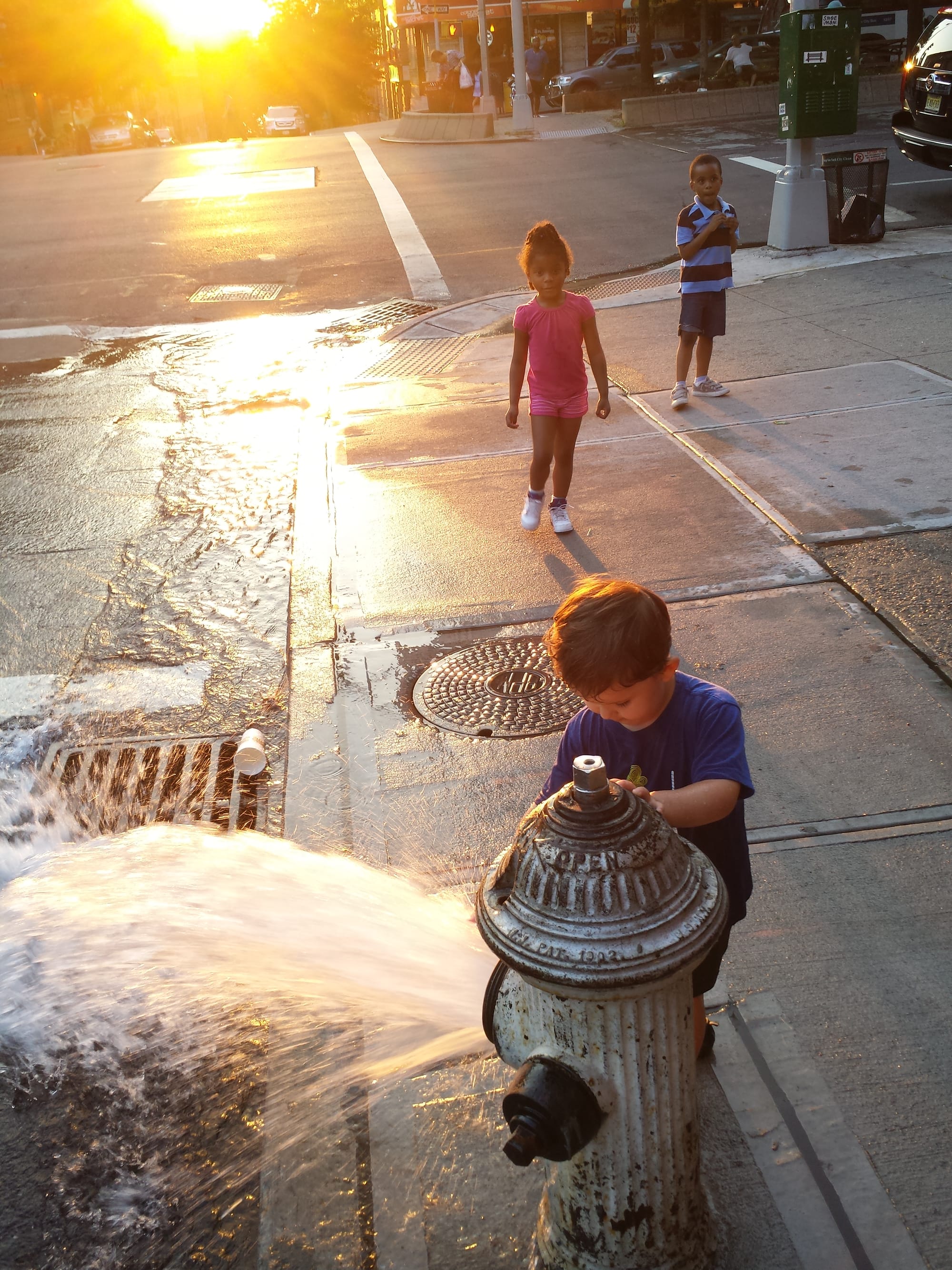 A child touching the water as it sprays out of a hydrant, with two other children standing close by as the sun sets.