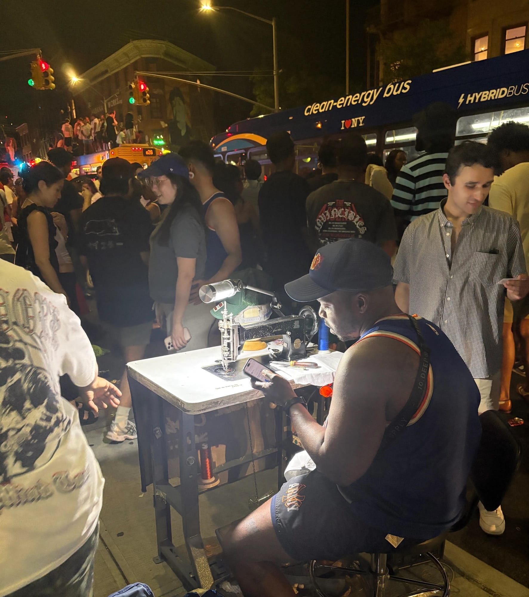 A man wearing a Knicks jersey sits at a Singer sewing machine outside at night surrounded by people.