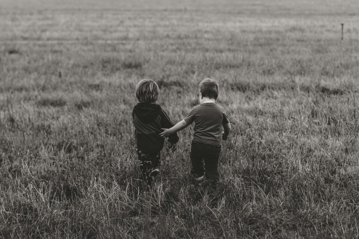 Black and white photo of two young children walking away from the camera through a grassy field. One child holds out their arm protectively toward the other. 