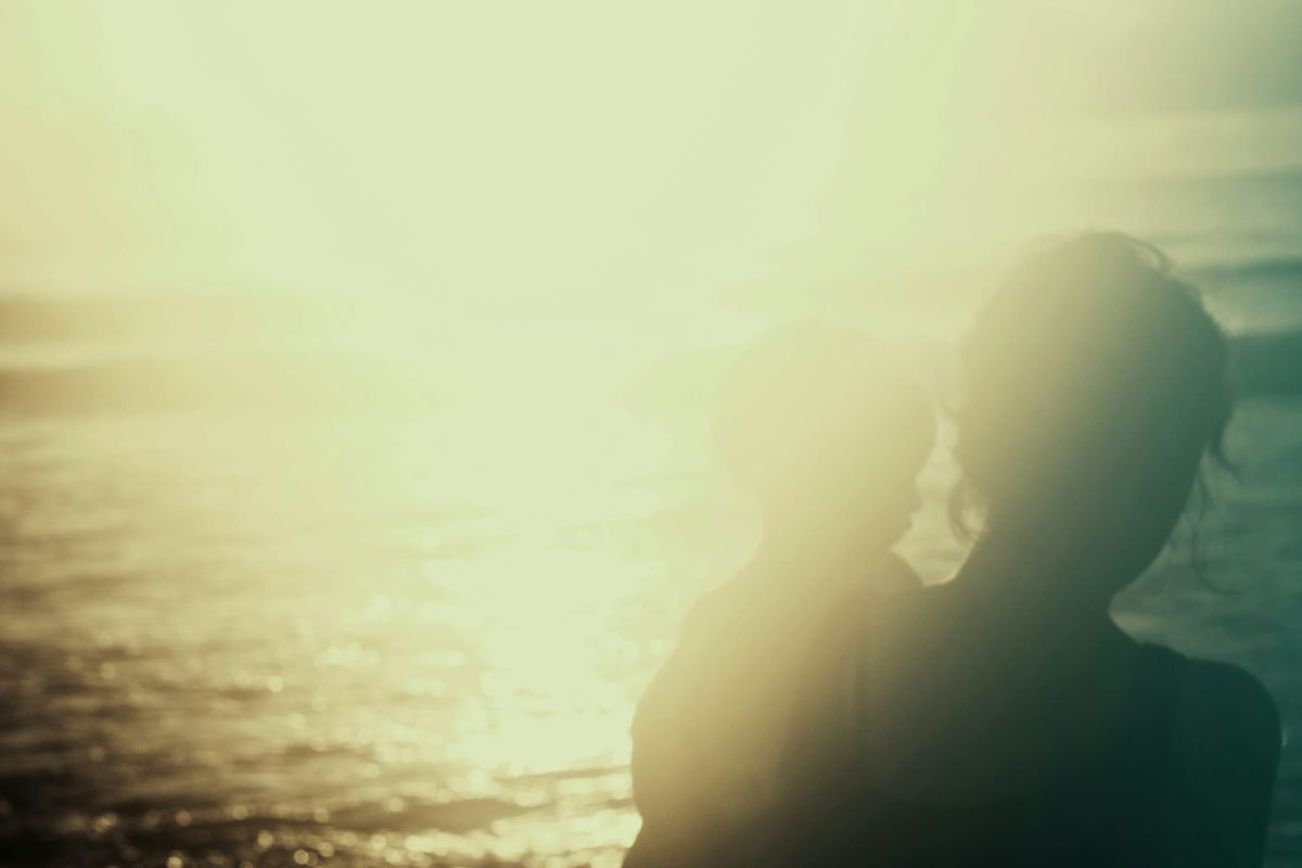 A hazy image of a woman carrying a small child. Water ripples in the sunlight beyond them.