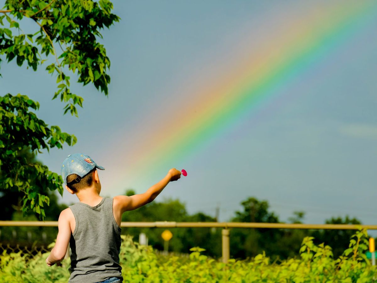 A child in a grey shirt and a blue baseball cap stands facing a rainbow and points into the distance, holding a red lolipop