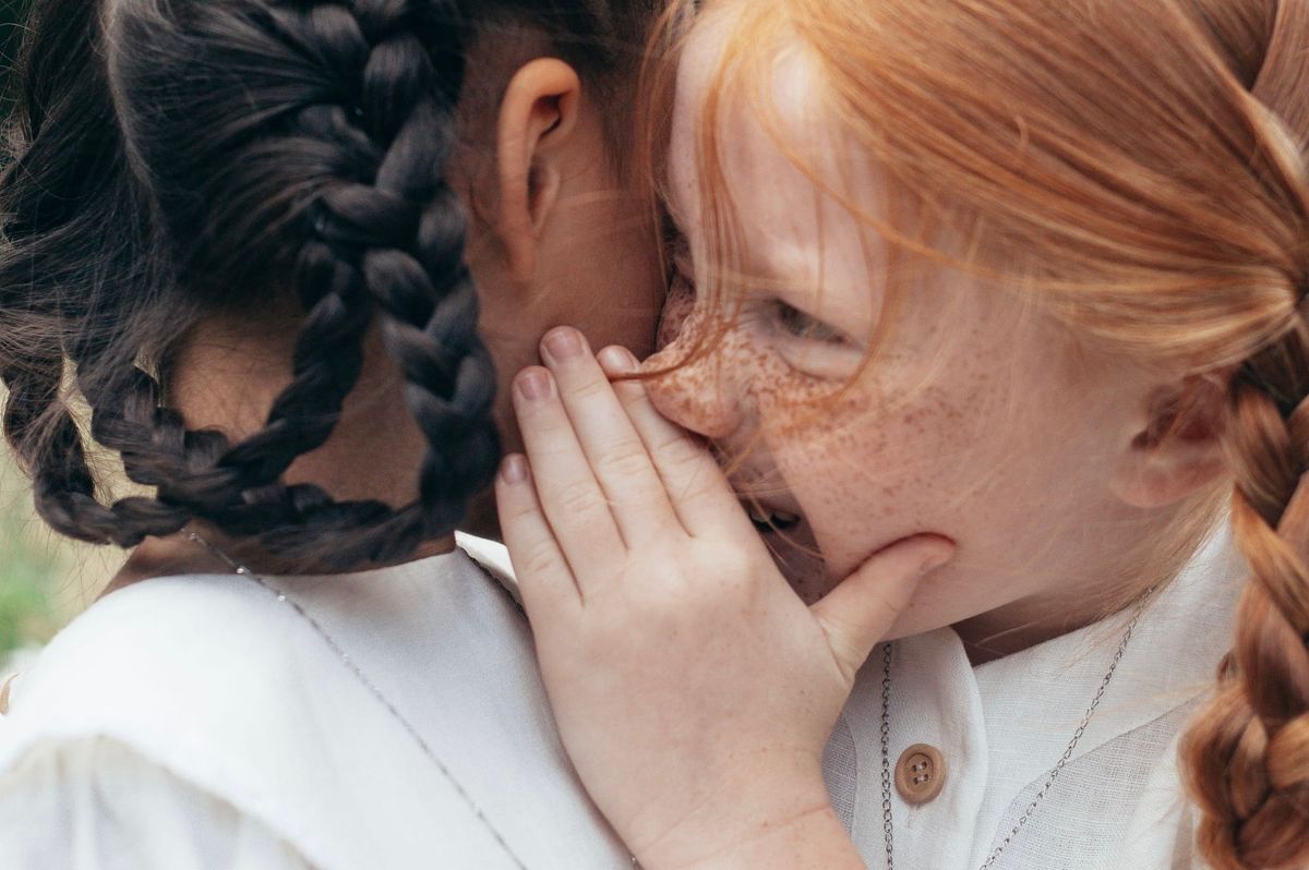 A child with red braids and freckles whispering in the ear of a child with dark brown braids.