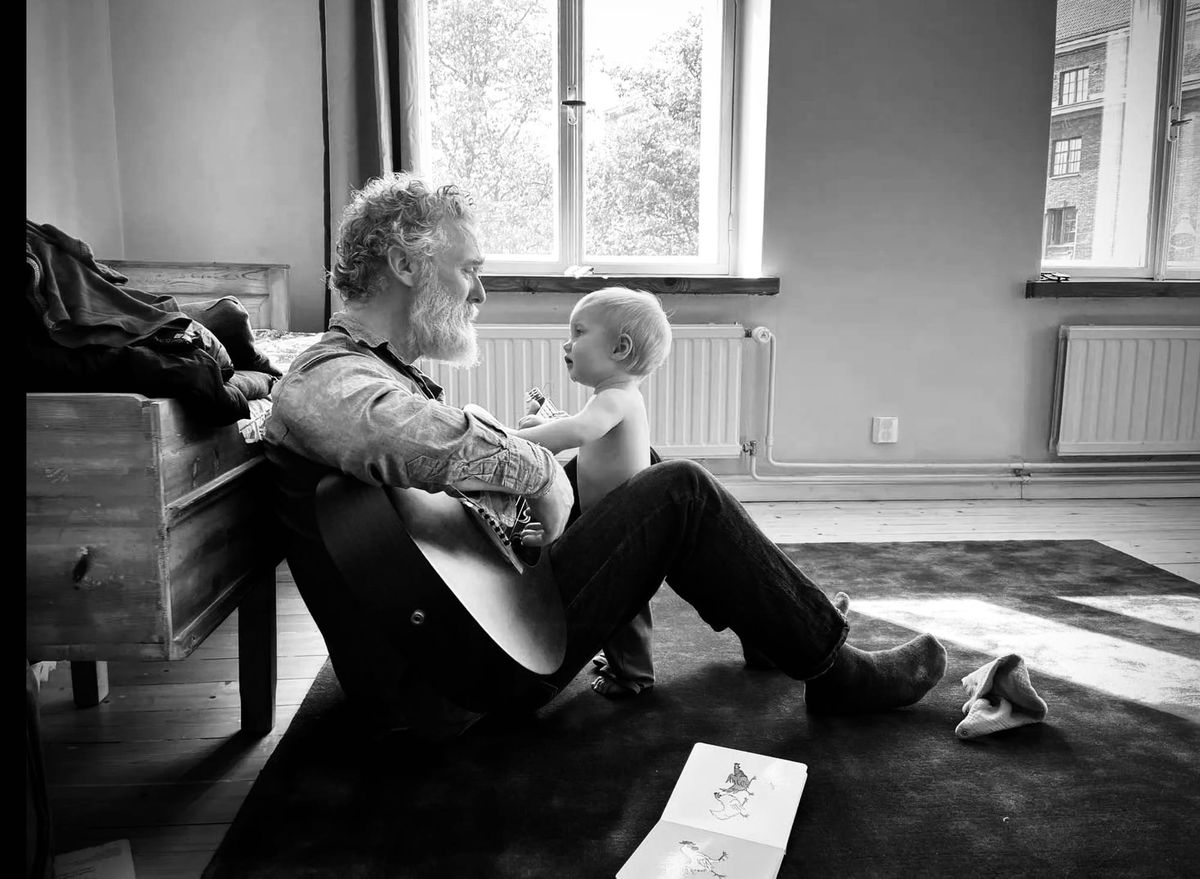 Black and white photo of Glen Hansard sitting on the floor with his guitar. His todler son is standing between his knees. They are looking at each other.