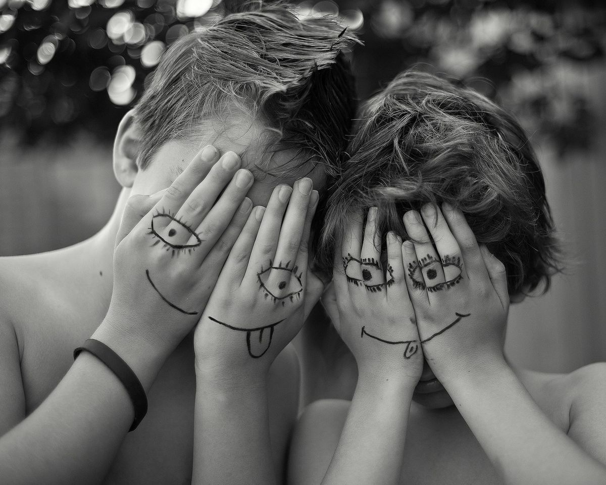 Black-and-white photo of two children covering their faces with their hands. Their hands have silly eyes and mouthes drawn on them to create the effect of masks. 