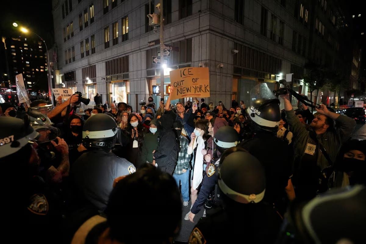 Police in riot gear stand in front of protesters at night in NYC. A man in the center holds a cardboard sign that reads "ICE Out of New York."