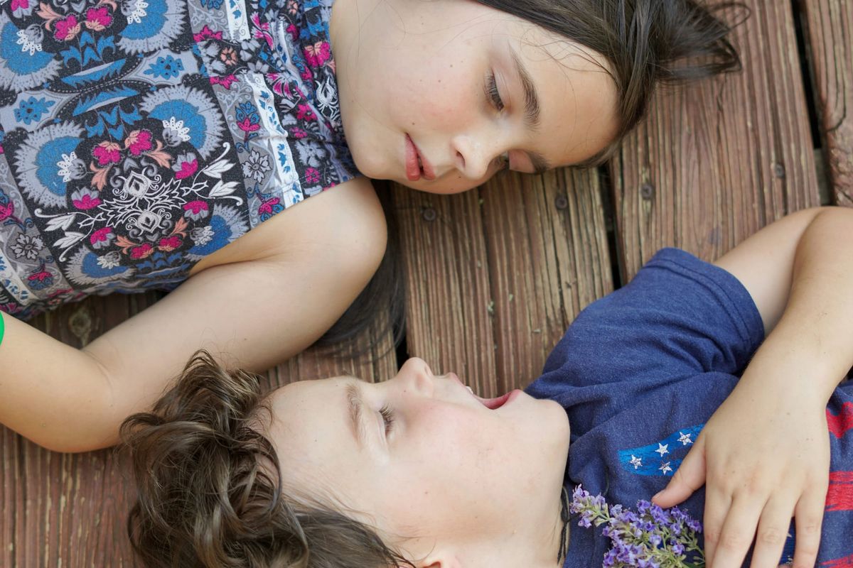 Two children lying on their backs, head-to-head, looking at eachother. The boy appears to be shouting while the girl looks at him with a steady gaze.