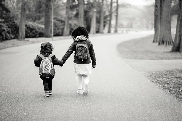 Photo of an older child holding a younger child's hand, viewed from behind, walking down a road