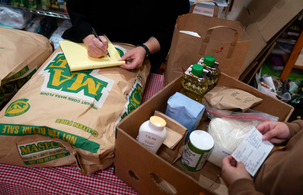 The hands of two volunteers packing boxes of groceries, holding lists of essential items