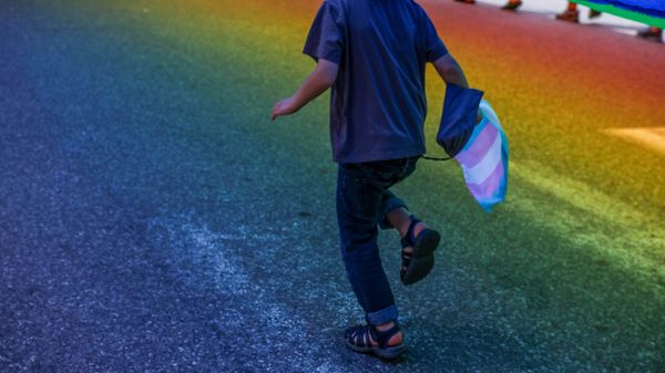 A child running through the colorful light cast by a rainbow flag above, holding a trans flag in one hand.