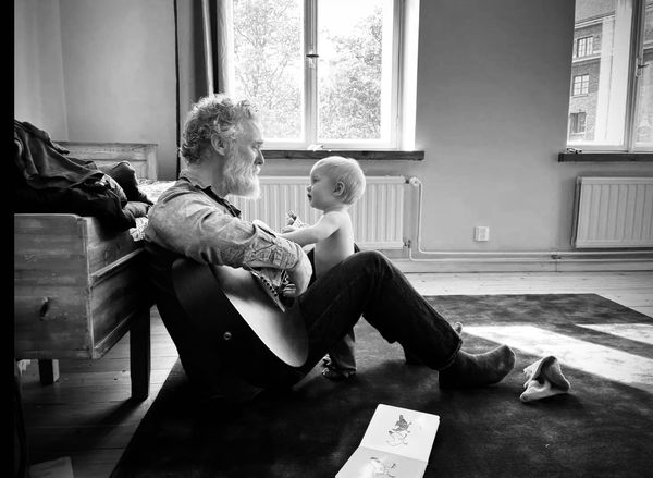 Black and white photo of Glen Hansard sitting on the floor with his guitar. His todler son is standing between his knees. They are looking at each other.