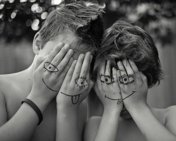 Black-and-white photo of two children covering their faces with their hands. Their hands have silly eyes and mouthes drawn on them to create the effect of masks. 
