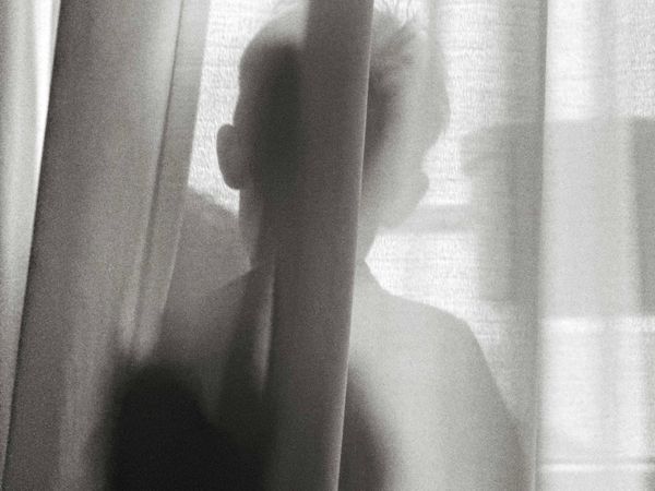 Black-and-white photo of the silhouette of a child sanding behind a curtain with their back to the camera., 