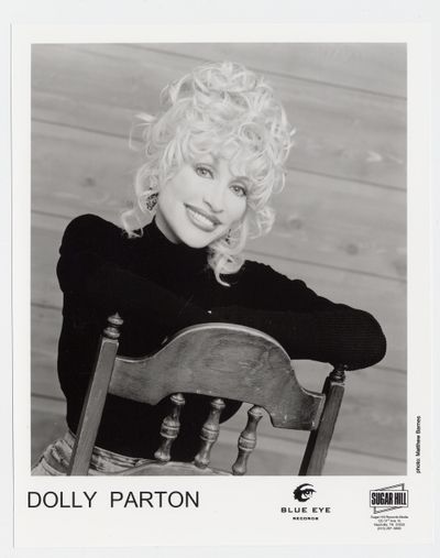 Black-and-white portrait of Dolly Parton smiling, with voluminous blonde curls, leaning on a wooden chair.